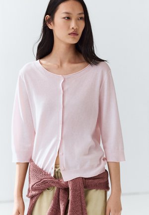 Woman with long dark hair wearing a light pink buttoned cardigan and a darker pink sweater tied around her waist.