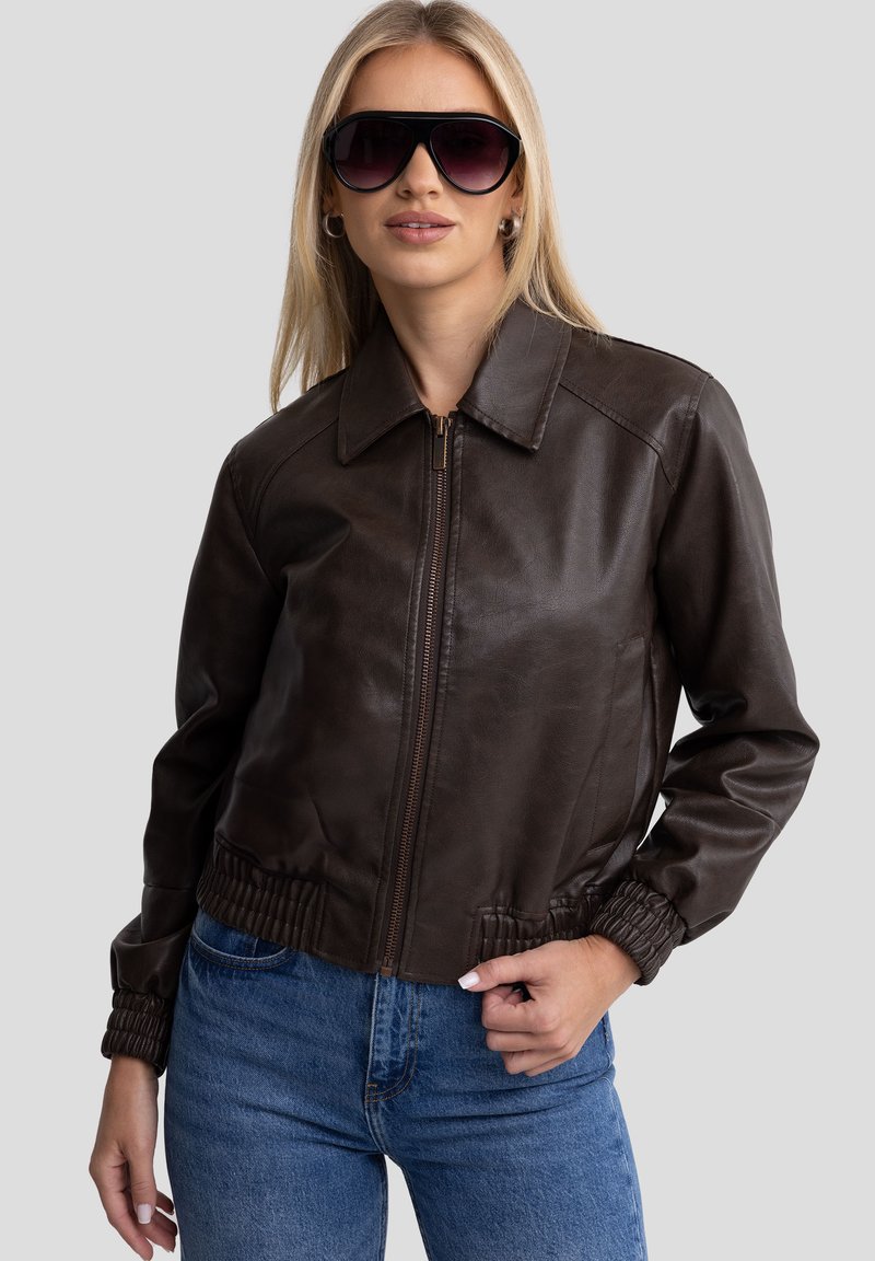Giacca bomber in pelle marrone con zip frontale, ampio colletto e polsini elasticizzati. Abbinata a jeans blu e occhiali da sole oversize scuri.