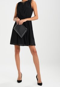 Svart ärmlös klänning med en figurnära midja och utsvängd kjol, prydd med spetsdetaljer vid halsringningen. Åtföljd av en svart kuvertväska med nitar.