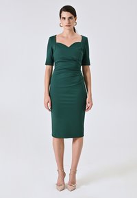 Grünes, tailliertes Kleid mit kurzen Ärmeln und herzförmigem Ausschnitt. Glatte Textur, knielang, kombiniert mit beigen, spitzen Absatzschuhen und Knöchelriemen.