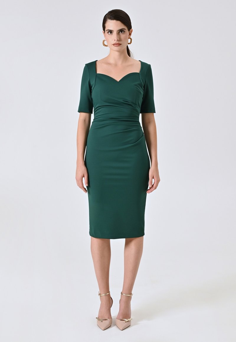 Grünes, tailliertes Kleid mit kurzen Ärmeln und herzförmigem Ausschnitt. Glatte Textur, knielang, kombiniert mit beigen, spitzen Absatzschuhen und Knöchelriemen.