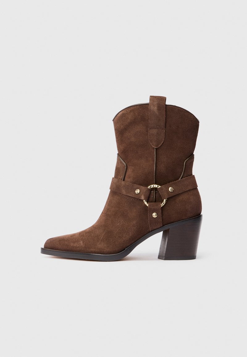 Bota cowboy de ante marrón con punta afilada, acentos en hardware en tono dorado y un resistente tacón marrón. Presenta un detalle de correa decorativa.
