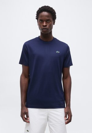 Muž nosí tmavomodrú košeľu s krátkym rukávom s malým zeleným logom krokodíla na hrudi a biele šortky.