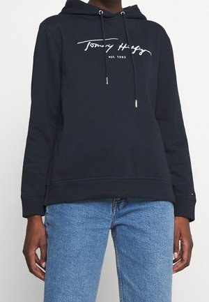 Personne portant un sweat à capuche noir Tommy Hilfiger avec cordons et un jean bleu, debout devant un fond clair uni.