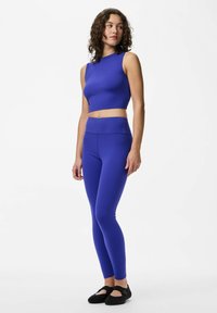 Ljubičasti crop top s visokim izrezom, uparen s odgovarajućim ljubičastim tajicama. Tkanina izgleda glatko i rastezljivo. Crne ravne cipele.