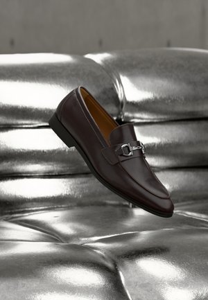 Mocassin en cuir marron foncé avec boucle en métal argenté reposant sur une surface matelassée argentée métallique brillante, incliné pour montrer les détails latéraux et supérieurs.