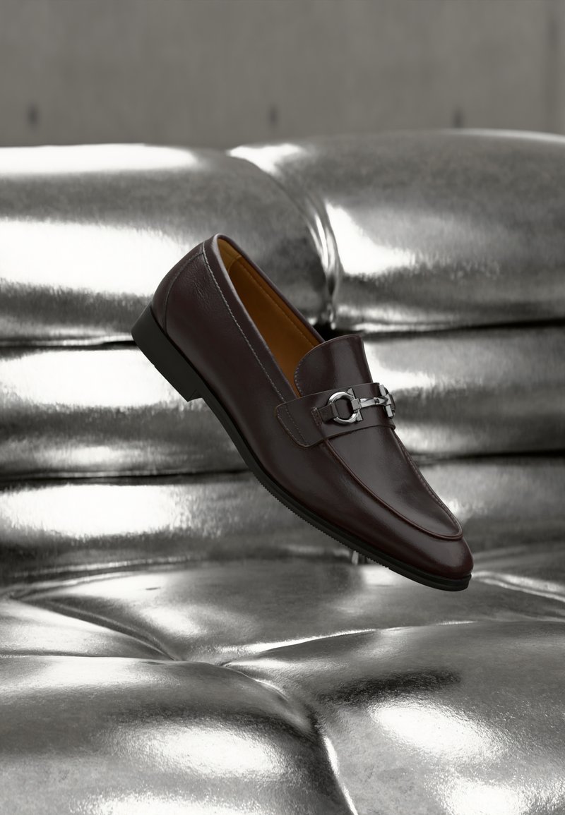 Mocassin en cuir marron foncé avec boucle en métal argenté reposant sur une surface matelassée argentée métallique brillante, incliné pour montrer les détails latéraux et supérieurs.