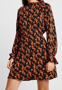 Svart klänning med ett orange blomstertryck, med hög halsringning och långa, puffiga ärmar med volangkantade manschetter. Kort fåll.