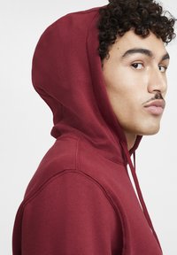 Bordeaux hoodie met een trekkoord, zachte textuur en een ontspannen pasvorm. Zijaanzicht benadrukt de prominente capuchon en stikdetails.