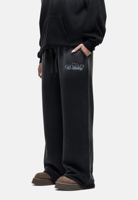 Zwarte sweatpants met een zachte textuur, voorzien van een geborduurd "GOOD" logo in het blauw op het linkerbeen, gecombineerd met bruine slippers.