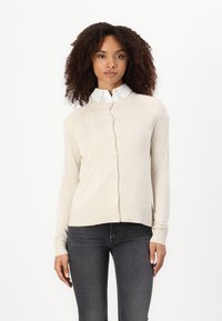 Beiger Cardigan mit Rundhalsausschnitt, Knopfverschluss und geripptem Saum. Über einem weißen Hemd mit Kragen getragen, kombiniert mit dunklen Jeans. Weiche Textur.