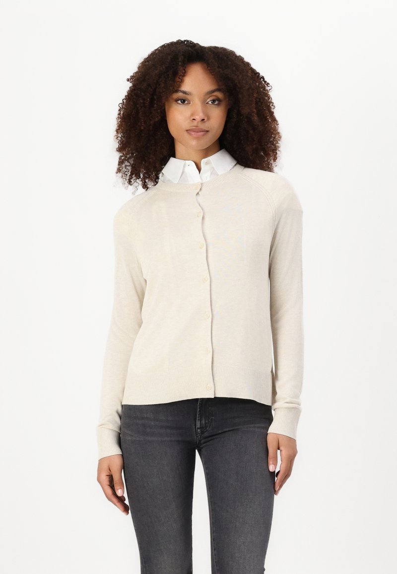 Beiger Cardigan mit Rundhalsausschnitt, Knopfverschluss und geripptem Saum. Über einem weißen Hemd mit Kragen getragen, kombiniert mit dunklen Jeans. Weiche Textur.