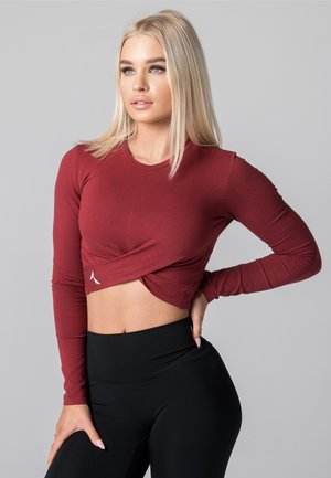 Blond naine poseerib ploomipunases pika varrukaga lühikeses ülemises osas ja kõrge vöökohtadega mustades retuusides, mille taustal on lihtne hall taust.