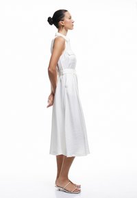 Vestido blanco sin mangas con cintura ceñida, que presenta una tela texturizada y un dobladillo a la altura de la rodilla. La modelo lleva sandalias blancas sencillas.