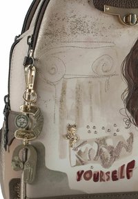 Leder-Rucksack mit beigem, strukturiertem Finish, geprägten Designs, Metallakzenten und einem dekorativen Katzenanhänger mit dem Spruch "Erkenne dich selbst".