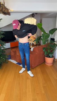 Mees, kes kannab punast mütsi, tõstab musta kapuutsi, et näidata paljast kõhtu siseruumides jõulupuu ja suure toalillepotiga lähedal.