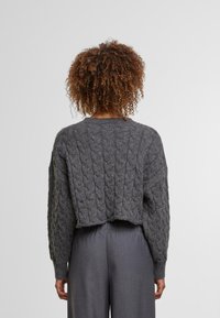 Henkilö, jolla on kiharainen hius, on pukeutunut lyhyeen, tummanharmaaseen palmikkoneuleeseen ja löysiin harmaisiin housuihin, selkä kameraan päin yksinkertaista taustaa vasten.