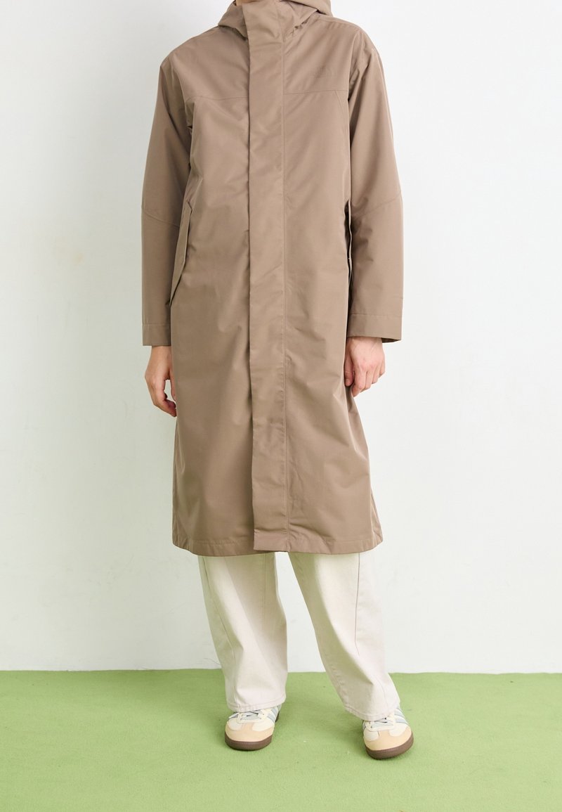 Personne portant un long imperméable beige à capuche sur un pantalon blanc ample et des baskets blanches, debout sur un sol vert contre un mur blanc.