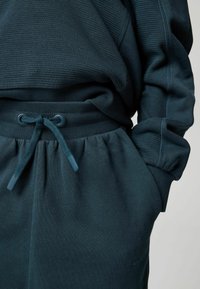 Tamsiai žalias svetimas su tekstūruotu audiniu ir derančiais džinsais. Yra raištelių juosmuo ir subtili prekės ženklo etiketė ant šlauko. Minkštas megztas audinys.