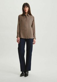 Chemise à manches longues avec un motif géométrique en noir, rouge et beige, assortie à un pantalon droit sombre et des bottes à talons noires.