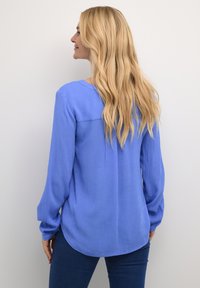 Camicia a maniche lunghe di un blu chiaro solido, tessuto testurizzato, orlo arrotondato, dettaglio della cucitura posteriore, abbinata a jeans blu scuro.
