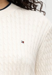 Kreminės spalvos megztinis su pynių raštu, apvalia apykakle ir tamsiai mėlyna detalė. Krūtinės srityje – mažas raudonos, baltos ir mėlynos spalvų logotipo lopas.