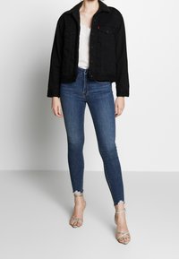 Svart denimjacka med krage av konstpäls, tillsammans med åtsittande blå jeans och sandaler med remmar i silver. Texturerad tyg med slitna detaljer.