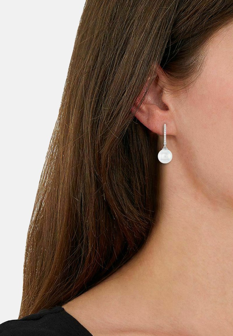 Mujer con cabello largo castaño que lleva un pendiente de plata con una perla blanca redonda.