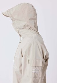 Giacca beige con cappuccio e colletto rialzato, cordino regolabile e tasca con zip sulla manica sinistra con logo ricamato bianco.