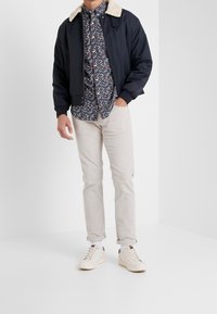 Giacca bomber navy con colletto in ecopelle crema, indossata sopra una camicia floreale multicolore, pantaloni beige chiaro e sneakers bianche.