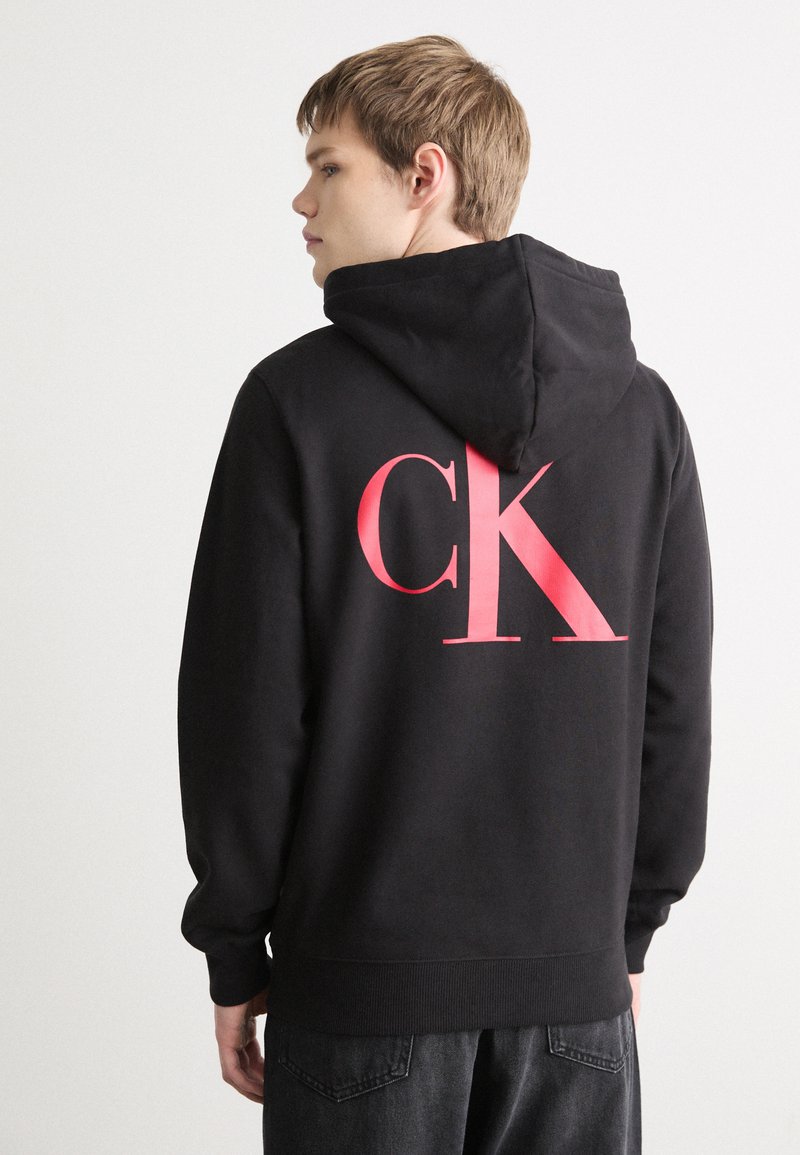 Melnā kapuce kokvilnas maisījumā, ar lielu rozā "CK" logotipu aizmugurē, kangaru kabatu un elastīgām aprocēm un apakšmalu.