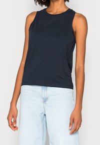 Kvinna med enärmad marinblå topp och ljusblå högmidjade jeans mot en enfärgad bakgrund.