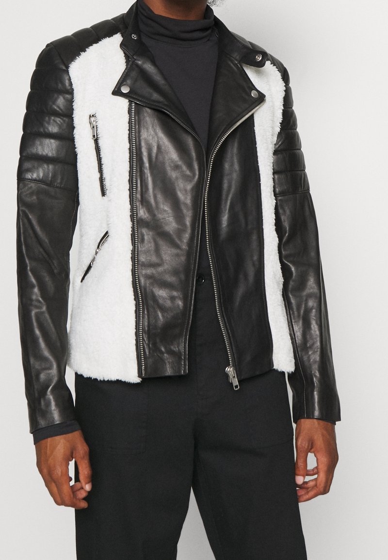 Chaqueta de cuero negra para hombre con paneles de piel blanca, cierre frontal con cremallera, bolsillos con cremallera y mangas acolchadas, usada sobre un top negro.