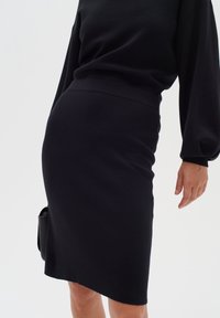 Femme portant une jupe noire ajustée longueur genou et un haut noir à manches longues bouffantes, tenant un sac à main noir.