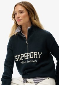 Kobieta z długimi blond włosami, nosząca granatowy sweter typu zip-up z napisem "SUPERDRY Athletic Essentials" na paskowanej koszuli z kołnierzykiem oraz białych spodniach.