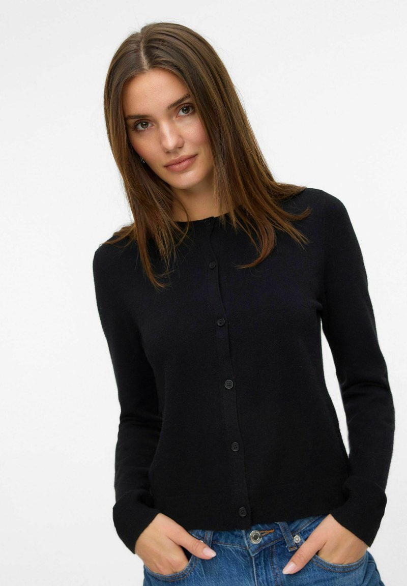 Cardigan noir en tricot avec des manches longues, un col rond et des boutons sur le devant. Présente une texture douce et une coupe ajustée. Associé à un jean bleu.