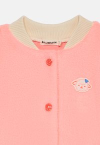 Rosa fleecejakke med en kremfarget ribbestrikket krage, glitrende rosa knapper og et sauemotiv på lappen. Merket leser "BILLIEBLUSH."