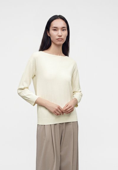 Femme aux longs cheveux noirs portant un haut crème à manches longues et un pantalon beige ample, debout devant un fond clair uni.