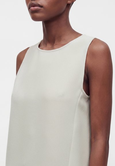 Femme portant un haut sans manches texturé beige clair, montrée des épaules au haut de la poitrine sur fond blanc uni.