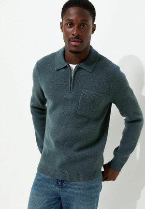 Giovane uomo che indossa un maglione grigio con collo a zip e tasca sul petto e jeans blu, in piedi davanti a uno sfondo bianco uniforme.