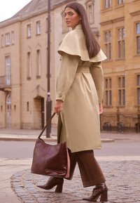 Trench coat khaki cu guler tip cape, talie cu curea și mansete cu nasturi. Pantaloni largi maro și o geantă mare burgundă. Botine cu toc texturate.