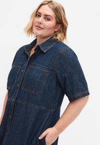 Tummansininen farkkuhenkseli, jossa on lyhyet hihat, kaksi napitettua rintataskua ja näkyvät ompeluakkentit. Taskut on suunniteltu käytännöllisyyttä varten.