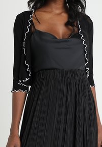 Svart cardigan med vita volanger, över en svart satin topp, tillsammans med en plisserad svart kjol. Har korta ärmar och en figurnära design.