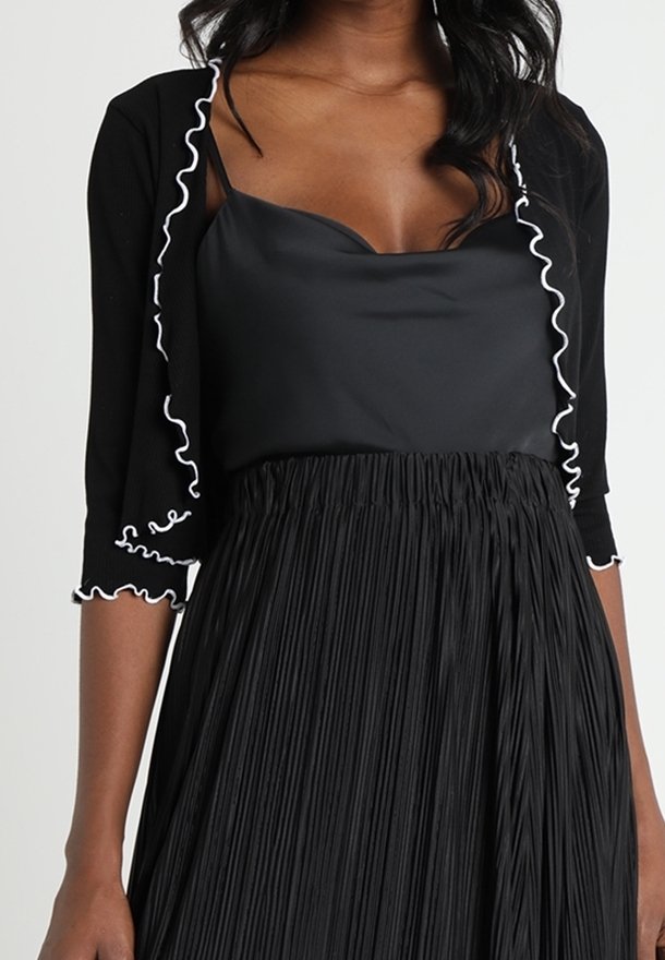 Svart cardigan med vita volanger, över en svart satin topp, tillsammans med en plisserad svart kjol. Har korta ärmar och en figurnära design.