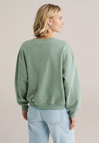 Oversized groene sweatshirts met een ronde hals, doorlopende schouders en geribbelde manchetten. Zachte textuur, gedragen met lichtblauwe spijkerbroeken.
