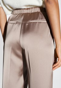Satijnen broek in lichtbruin met een elastische tailleband en zijzakken. De stof heeft een gladde textuur en een subtiele glans.