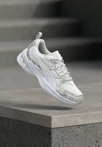 Άσπρο και ασημί αθλητικό παπούτσι κρεμασμένο πάνω από μια σκυρόδετη προεξοχή με θολές σκάλες στο παρασκήνιο.