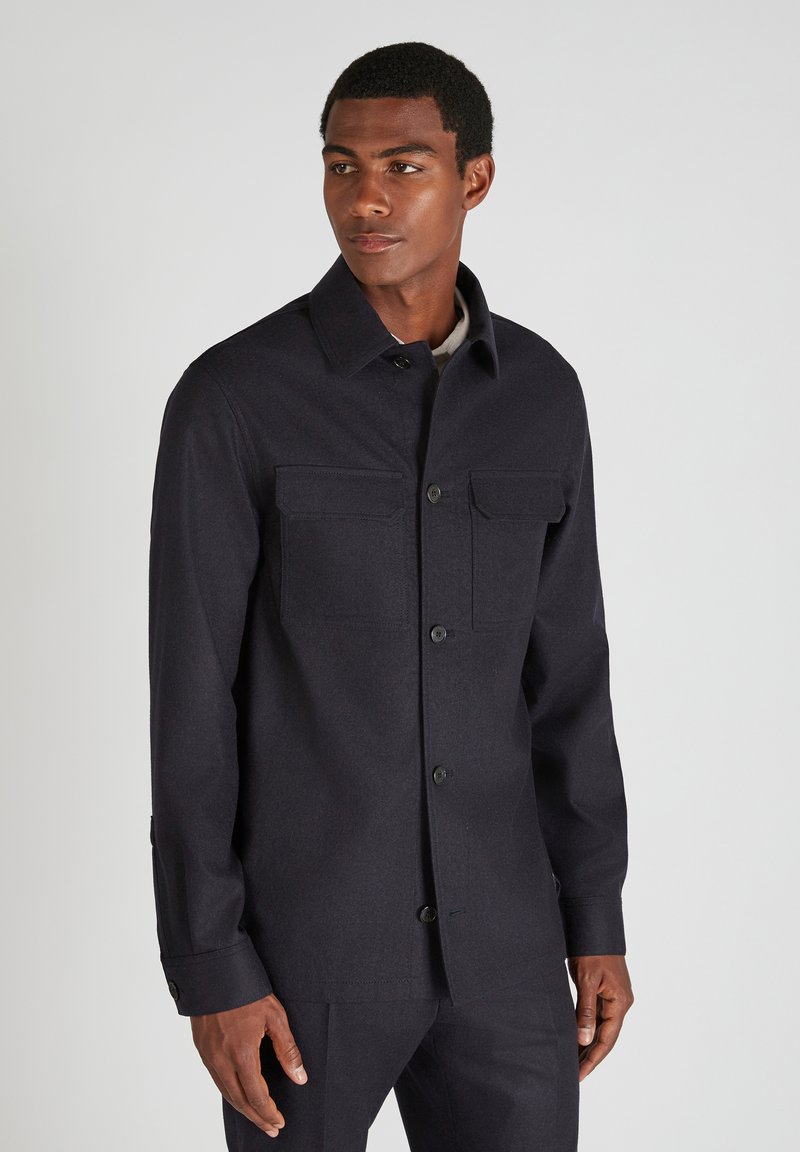 Giacca di lana blu scuro con collo, due tasche sul petto e chiusura a bottoni. Presenta un tessuto testurizzato e una silhouette aderente.