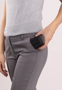 Zwarte leren portemonnee met een gestructureerde afwerking, gedeeltelijk zichtbaar in een grijze broekzak. De broek is maatwerk en heeft een fijne uitstraling.