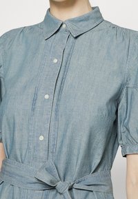 Světle modré denimové šaty s krátkými rukávy, knoflíkovým zapínáním vpředu, špičatým límcem a látkovým páskem uvázaným do mašle v pase.